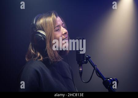 Eine niedliche junge Frau mit Kopfhörern singt in einem dunklen Raum in ein Mikrofon. Stockfoto