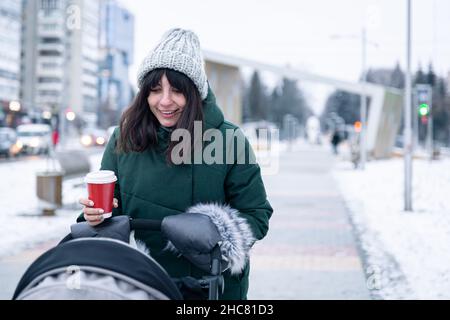 Stilvolle junge Mutter mit einer Tasse Kaffee auf einem Spaziergang mit einem Kinderwagen im Winter. Stockfoto