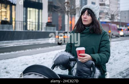 Stilvolle junge Mutter mit einer Tasse Kaffee auf einem Spaziergang mit einem Kinderwagen im Winter. Stockfoto