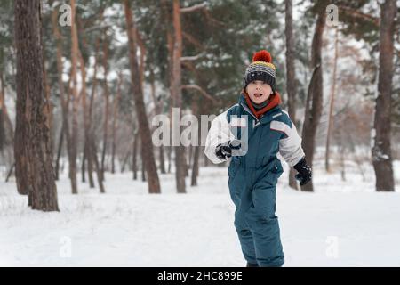 Der kleine Junge läuft fröhlich durch den Schnee. Kind spielt im Winterwald auf großen Tannenbäumen Hintergrund Stockfoto