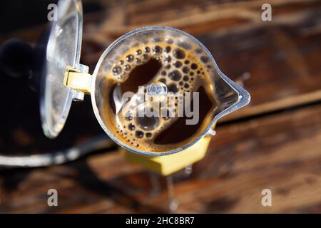 Gelbe Moka Kaffeedose auf dem Herd auf dem Tisch mit offener Abdeckung und braunem Heißgetränk im Inneren. Old Style Kaffee Vintage Topf Outdoor Camping Stockfoto