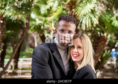 Schwarzer Mann und blonde Frau blicken auf die Kamera, während sie während eines romantischen Dates an einem sonnigen Tag im exotischen Park gegen Palmen stehen Stockfoto
