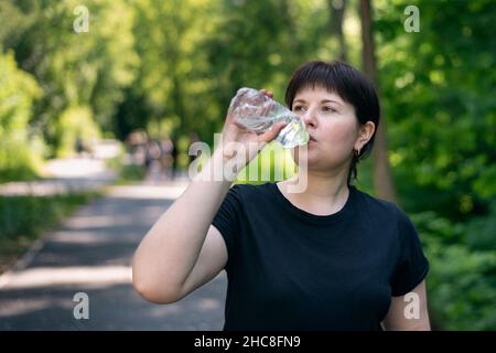 Die junge Frau trinkt Wasser, während sie im Park joggt. Trinkregime während des Sports Stockfoto