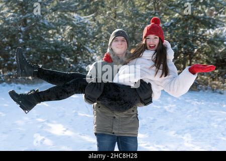 Lächelnder junger Mann hält seine Geliebte in den Armen, während er durch den Winterwald geht. Verliebte Paare. Stockfoto