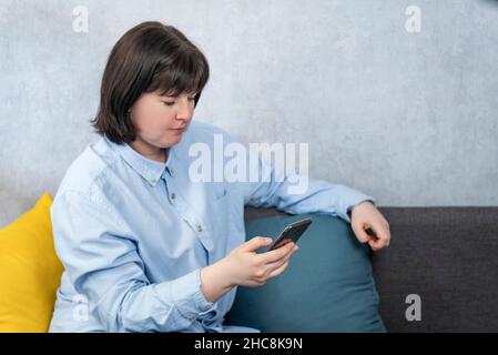 Eine dicke junge Frau im strengen Hemd sitzt auf der Couch mit dem Telefon in den Händen. Stockfoto
