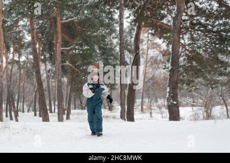 Der kleine Junge läuft fröhlich durch den Schnee. Kind spielt im Winterwald auf großen Tannenbäumen Hintergrund Stockfoto