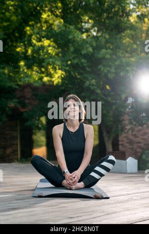 Das Porträt einer jungen Frau in Sportbekleidung sitzt draußen auf einer Yogamatte. Yoga am Morgen im Freien üben Stockfoto