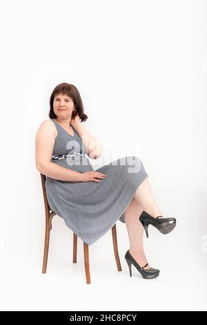 Junge glückliche Frau, die im Kleid übergewichtig ist, sitzt auf einem Stuhl auf weißem Hintergrund. XXL-Modell Stockfoto