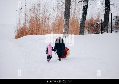 Entferntes Foto von weiblichem Kind und Mutter mit Schlitten, die beide in die Kamera blicken und im Schnee im Wald zur Kamera gehen. Erstaunlicher Hintergrund voller Stockfoto