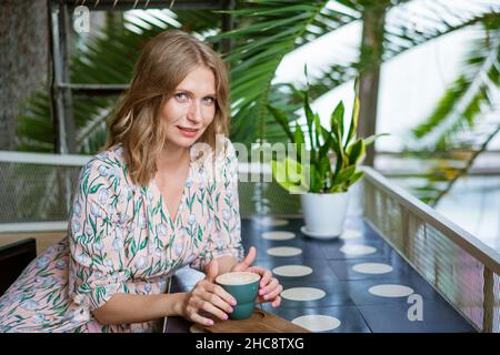 Fröhliche lächelnde Frau mittleren Alters, die in der Cafeteria sitzt und einen Kaffeebecher zur Seite hält. Eine Frau mittleren Alters trinkt Tee und denkt nach. Entspannung und Besinnung bei einer Tasse Kaffee. Stockfoto
