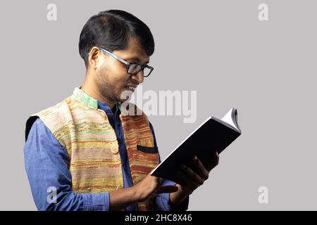 Ein junger indischer Mann in ethnischer Kleidung liest Buch auf grauem Hintergrund Stockfoto