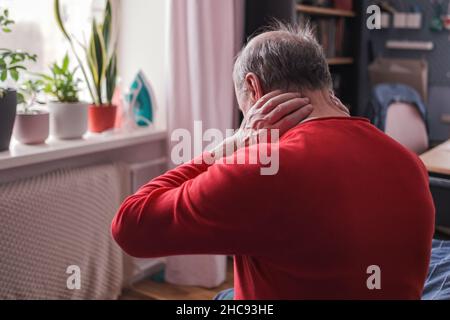 Frustrierter älterer Mann in rotem Pullover, der die Hand auf dem Nacken hält und auf dem Sofa sitzt Stockfoto