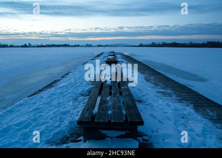Holzbank auf einem schneebedeckten Pier und einem gefrorenen See nach Sonnenuntergang, Zoltance, Polen Stockfoto