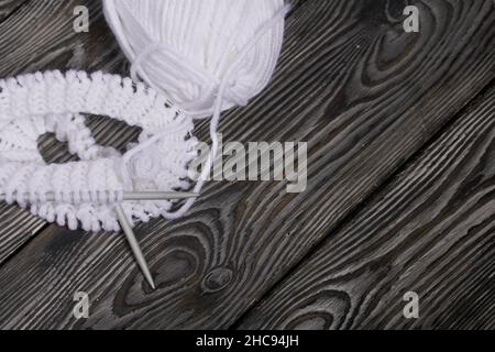 Stricken mit Nadeln. Ein weißer Faden und ein Fragment eines Strickprodukts. Auf schwarzen Kiefernbrettern. Nahaufnahme. Stockfoto