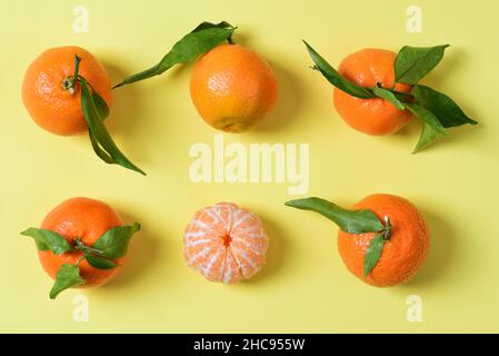 Sechs Mandarinen liegen nebeneinander auf gelbem Grund. Auf den Mandarinen befinden sich noch einige Blätter. Eine Mandarine wird geschält. Stockfoto