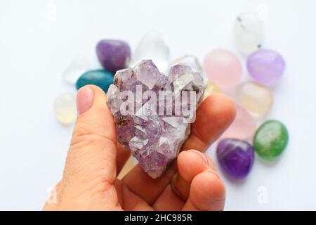 Wunderschöne purpurrote Kristalle aus Amethyst in der Hand. Druse Amethyst in Herzform auf dem Hintergrund anderer Edelsteine. Stockfoto