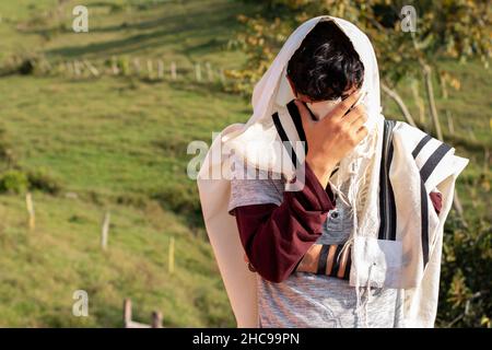 Jude trägt Tallit und Tefillin oder Mylakterie und betet in der Natur, während er sein Gesicht mit den Händen bedeckt. Stockfoto