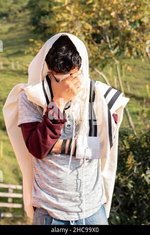 Vertikales Foto eines Juden, der in der Natur Tallit und Tefillin oder Mylakterie trägt und sein Gesicht mit den Händen bedeckt. Stockfoto