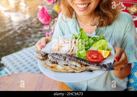 Eine glückliche Frau hält in einem Fischrestaurant einen Teller mit frisch gebackenen Regenbogenforellen Stockfoto