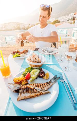 Fröhlicher Mann mit gegrilltem Seebassfisch und anderen Gerichten in einem Fischrestaurant. Das Konzept der mediterranen Ernährung und des epicure Lifestyles Stockfoto