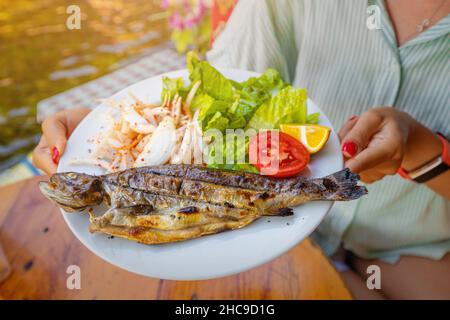 Eine glückliche Frau hält in einem Fischrestaurant einen Teller mit frisch gebackenen Regenbogenforellen Stockfoto