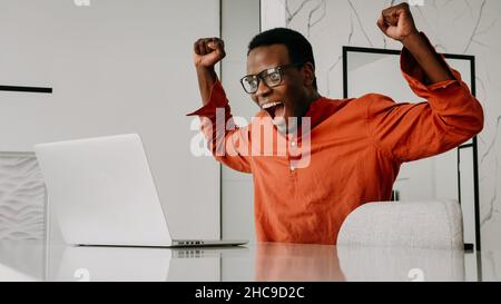 Fröhlicher afro-amerikanischer Freiberufler freut sich, von zu Hause aus an einem Laptop zu arbeiten, fröhlicher schwarzer Mann mit Brille und orangefarbenem Hemd hob seine Hände hoch und schaute auf den Computer oder sah die gute Nachricht Stockfoto