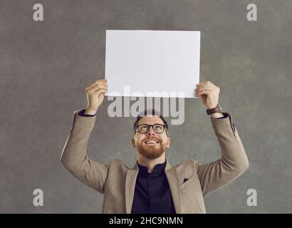 Ein junger Mann mit Brille und rotem Bart hält ein weißes, blank geteiltes Stück Papier über seinen Kopf. Stockfoto
