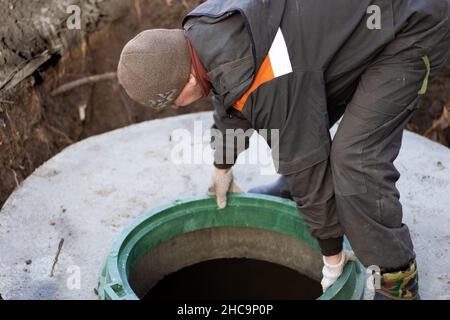 Ein Mann installiert einen Kanalkanal auf einen Klärgruben aus Betonringen. Bau von Kanalnetzen im Dorf. Stockfoto