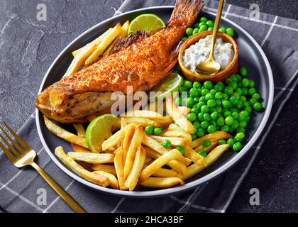Gebratener Seesockel auf einem Teller mit Kartoffelchips, gekochten grünen Erbsen, Tartarsauce und Limettenscheiben, horizontale Ansicht von oben, Nahaufnahme Stockfoto