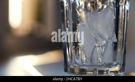 Eisstücke in ein transparentes Glas werfen und einen Cocktail zubereiten. Nahaufnahme eines Kristallglases, das auf dem Tisch steht und mit kaltem Eis auf dem verschwommenen Rücken gefüllt ist Stockfoto