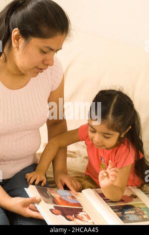 3-jähriges Mädchen zu Hause mit Mutter, die sich Familienfotos im Album Talking and Interaging ansieht Stockfoto