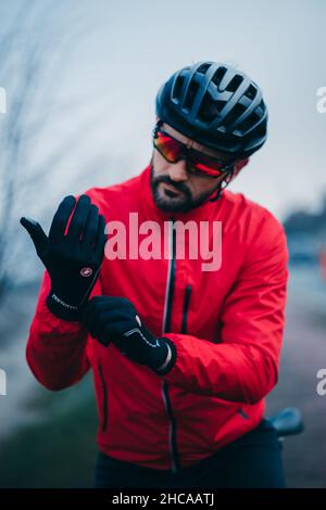 Junger Radsportler in roter Sportkleidung, der seine Castelli Perfetto Radsport-Winterhandschuhe in Schwarz anpasst Stockfoto