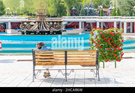 Rückansicht einer Frau, die auf der Bank vor dem Brunnen im First President's Park sitzt - einem städtischen Wahrzeichen in Almaty, Kasachstan Stockfoto