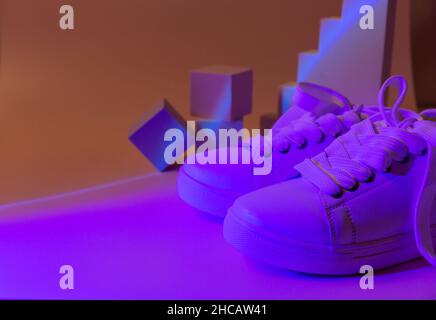 Trendige Mode weiße Sneakers auf Geometrie Podium auf abstrakt hellen Hintergrund. Neonlichter auf Freizeitschuhen. Violettes und orangefarbenes Gradientenlicht. Minimal Stockfoto