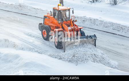 Großer orangefarbener Traktor reinigt Schnee von der Straße und lädt ihn in den LKW. Reinigung und Reinigung der Straßen in der Stadt vom Schnee im Winter Stockfoto