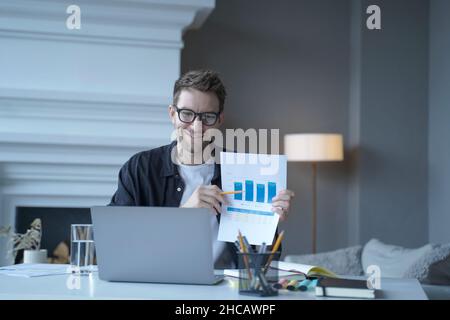 Junger lächelnder deutscher Geschäftsmann, der während eines Internettreffens mit Grafiken auf das Dokument zeigt Stockfoto