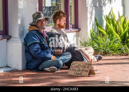 Zwei Bettler sitzen auf dem Bürgersteig mit einem Pappschild und bitten um Weihnachtsstimmung. Stockfoto