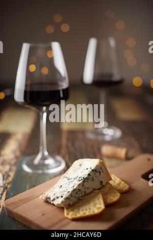 Stillleben von zwei Gläsern Wein, Käse mit blauem Schimmel und Crackern auf einem Holzbrett vor dem Hintergrund einer Neujahrskirlande. Vorderansicht. Stockfoto