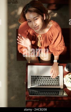Lächelnde junge Frau, die auf dem Laptop codiert, die Kamera anschaut und mit der Hand winkt Stockfoto