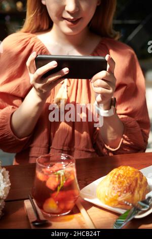 Junge Frau fotografierte Fruchtcocktails und Gebäck, das sie zum Mittagessen im Café bestellte Stockfoto