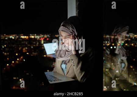Junge gestresste muslimische Geschäftsfrau, die beim Aufstehen am Fenster auf den Laptop-Bildschirm schaut und den Tempel berührt Stockfoto