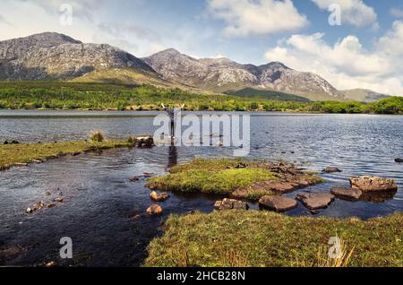 Wunderschöne Landschaft wilth Person springen in der Luft auf dem Felsen am lough inagh mit Bergen im Hintergrund im Connemara Nationalpark, Grafschaft Galway, Irland Stockfoto