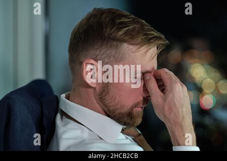 Seitenansicht des Gesichts eines jungen, müden Geschäftsmannes, der am Arbeitsplatz im Büro sitzt und nachts Überstunden arbeitet Stockfoto