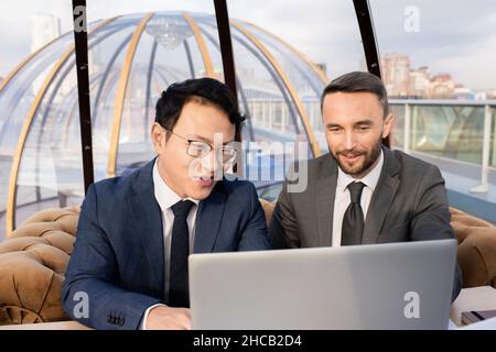 Zwei erfolgreiche Geschäftsleute in Anzügen, die beim Treffen im Café auf den Laptop-Bildschirm schauen und Daten diskutieren Stockfoto