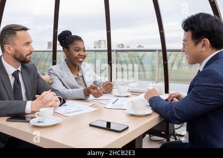 Glückliche junge afrikanische Ökonomin in elegantem Anzug, die mit chinesischen Kollegen beim Geschäftstreffen im Café interagiert Stockfoto