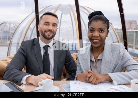 Zwei glückliche junge interkulturelle Ökonomen sitzen bei einem Treffen am Tisch im Café und blicken lächelnd auf die Kamera Stockfoto