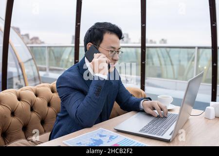 Junger, beschäftigter chinesischer Broker, der auf dem Mobiltelefon spricht, während er vor dem Laptop am Tisch sitzt und auf das Display schaut Stockfoto