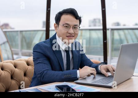 Fröhlicher junger chinesischer Geschäftsmann in formeller Kleidung und Brillen, der vor dem Laptop arbeitet, während er im Café am Tisch sitzt Stockfoto
