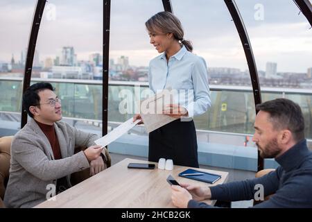 Fröhliche junge Kellnerin, die einem interkulturellen Geschäftsmann, der im Café oder Restaurant am Tisch sitzt, ein Menü übergab Stockfoto