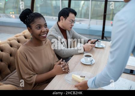 Fröhliche junge afrikanische Frau in eleganter Casualwear, die Kellnerin ansieht, die im Café einen Teller mit Käsekuchen auf den Tisch legt Stockfoto